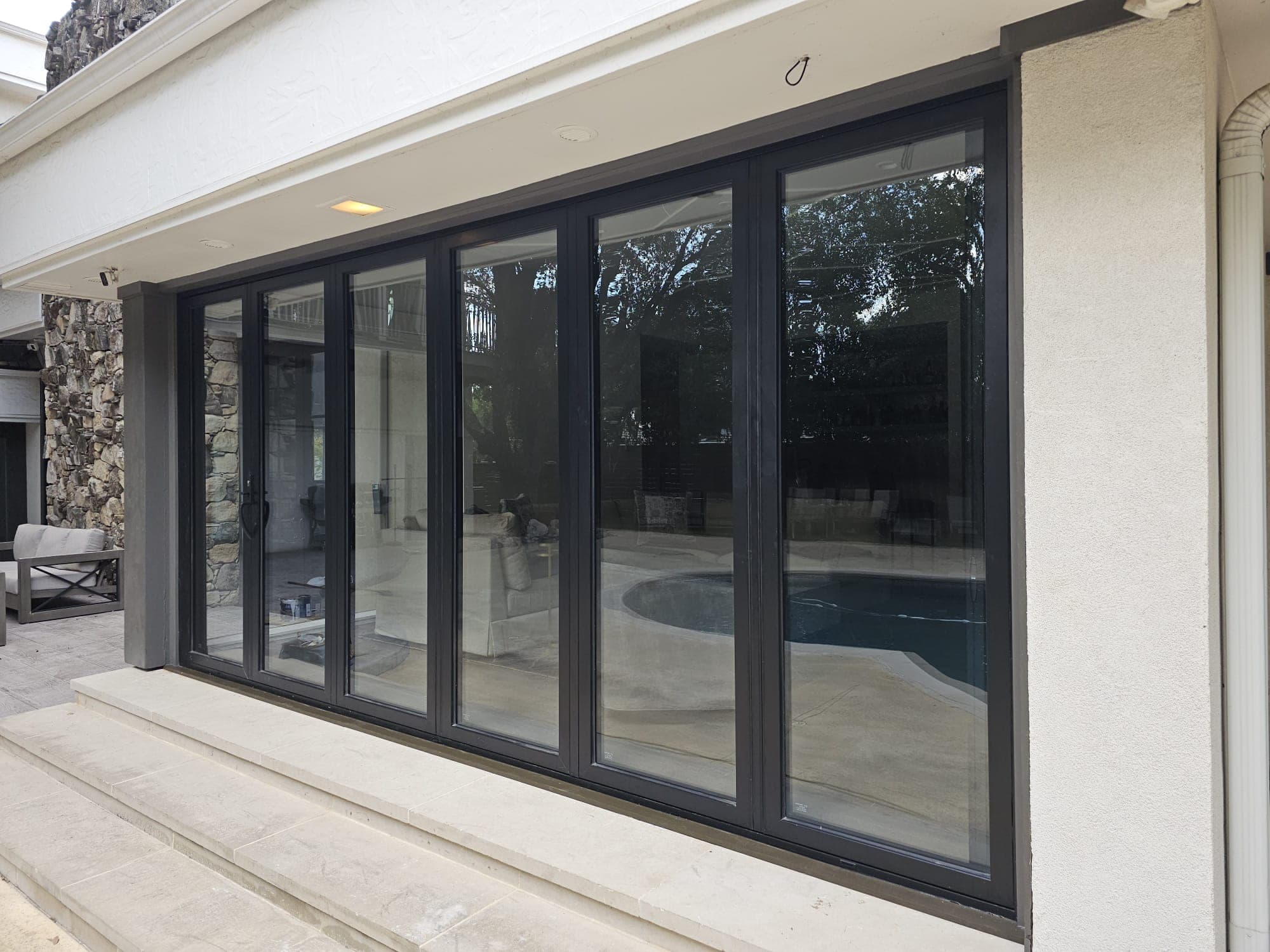 Five-panel black sliding door system set into a stucco wall beside a stone chimney, pool reflected in the glass
