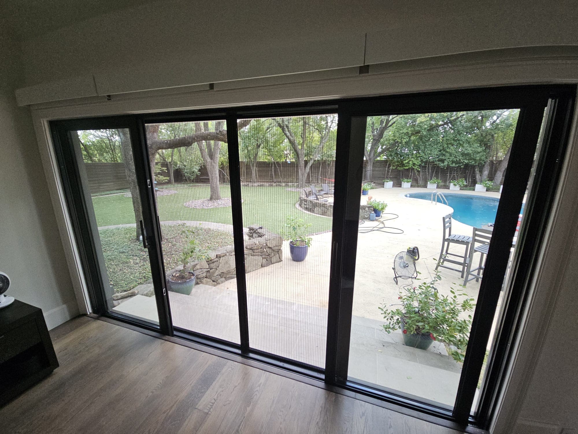 Interior view looking out through a black-framed sliding door system to a lawn, pool and mature oaks