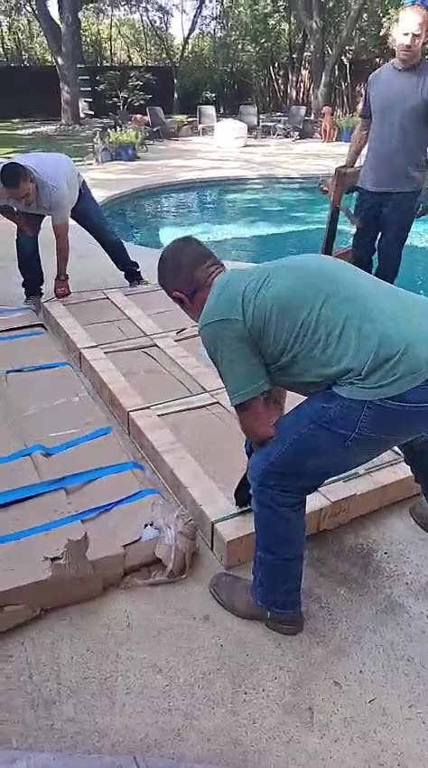 Three crew members lifting a large crated door panel across a pool deck